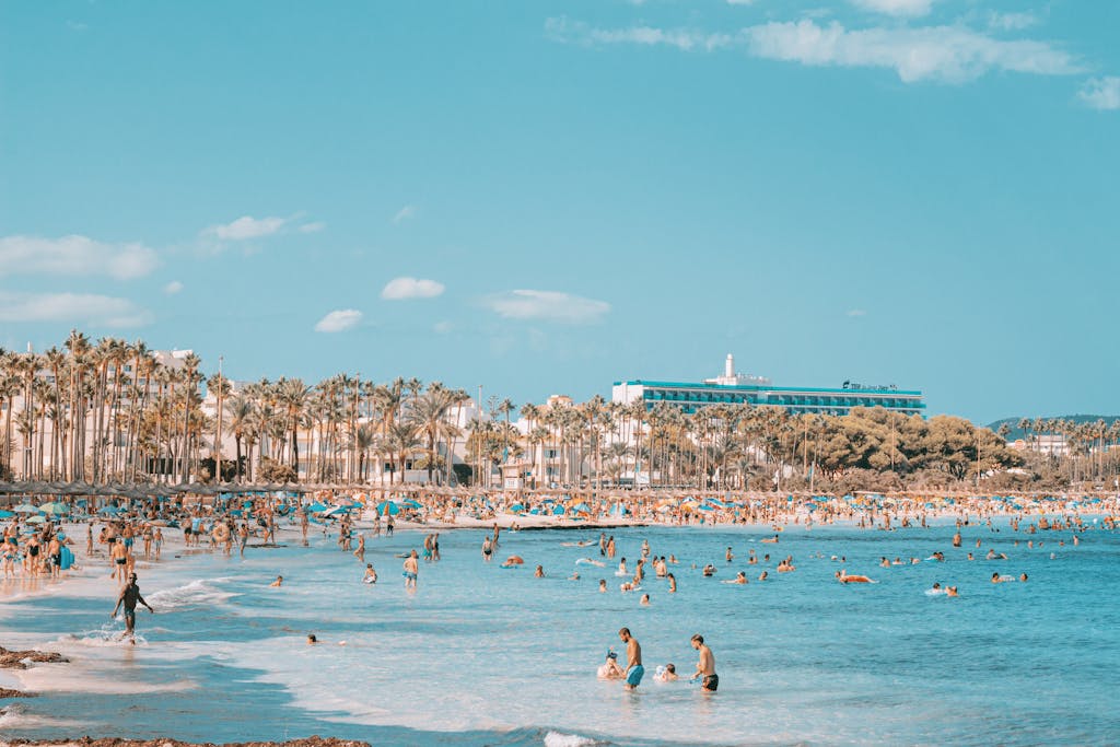 A vibrant summer day at the crowded beach in Palma de Mallorca, Spain, with clear blue waters and palm trees.