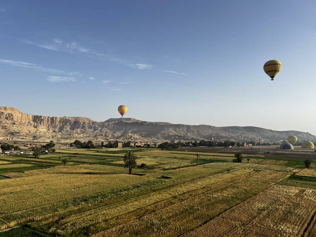 Green fields of Luxor with the mountains in the background and hot air balloons flying in the sky. 