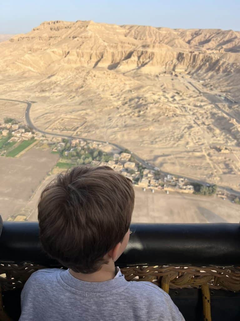 Child looking over the edge of the ballon basket while flying over the desert on the west bank of Luxor with Hatshepsut temple in the background. 