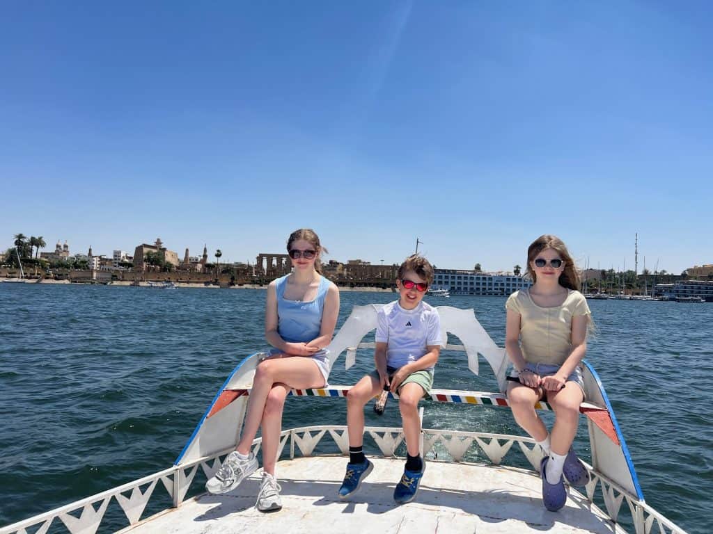Three children sitting on the top of a taxi boat crossing the river Nile in Luxor. Luxor temple is visible in the background on the far bank of the river.