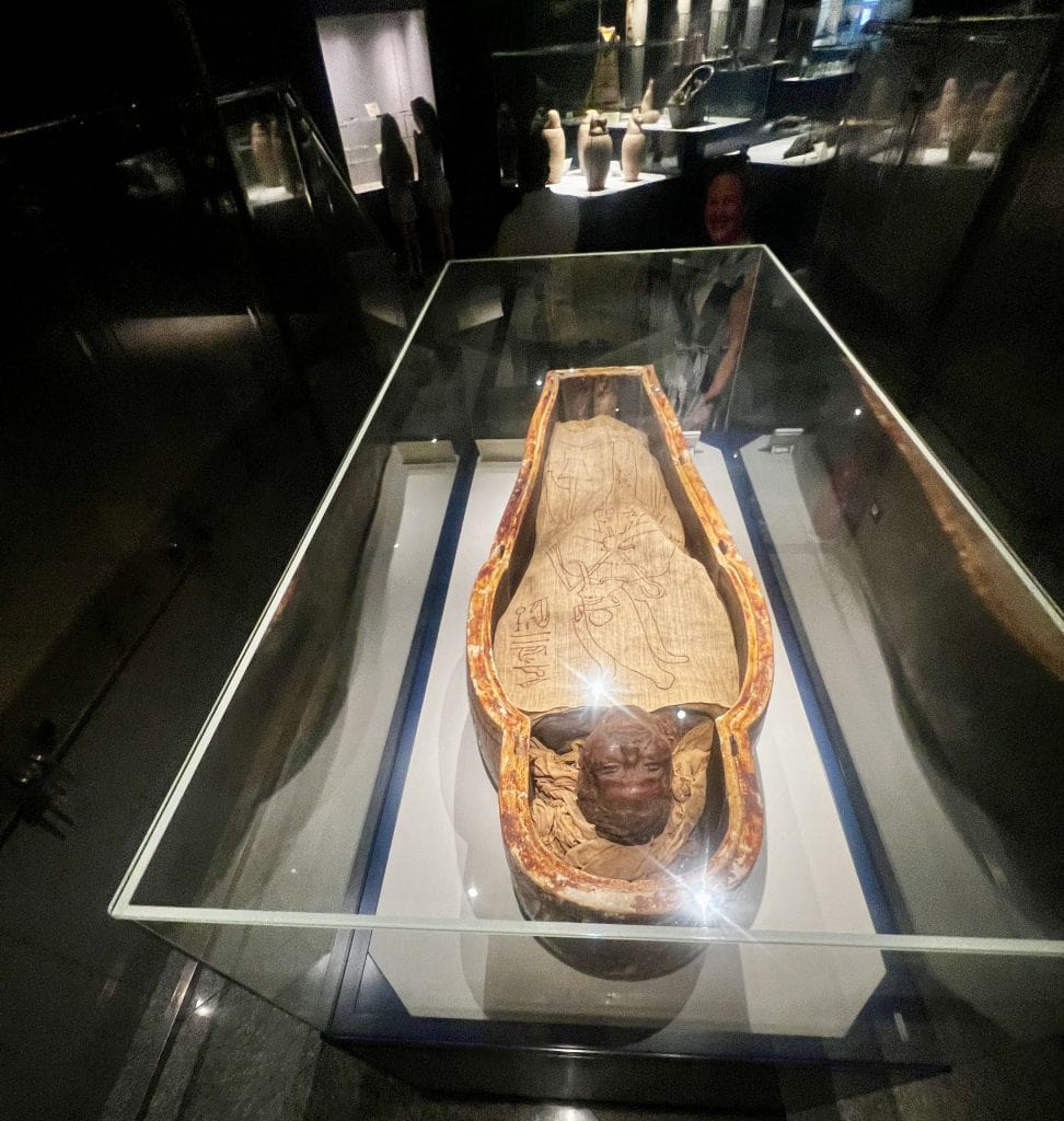 The inside of the Mummification museum in Luxor. In the foreground is a mummy in a glass display cabinet, with other exhibits visible in the background.