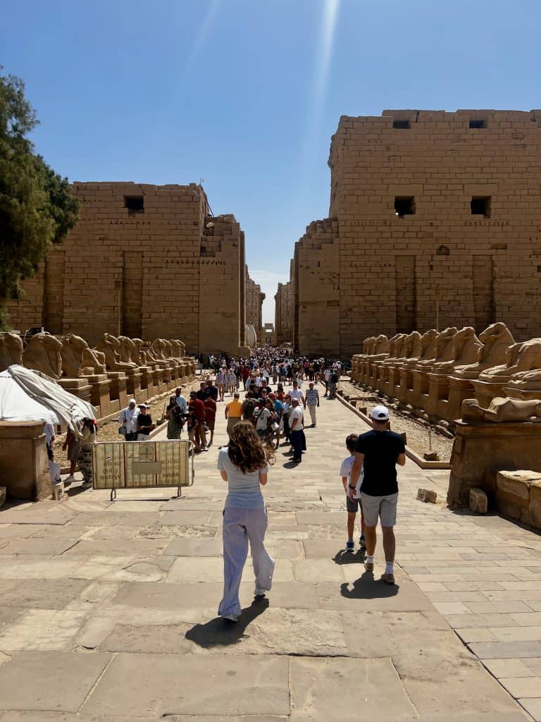 Adam, Chloe and Benjamin walking towards the main section of the Karnak Temple complex in Luxor. Behind can be seen a wide walkway lined with statues of rams on plinths. Behind these are the tall walls of the complex, with crowds of people seen walking around within them.