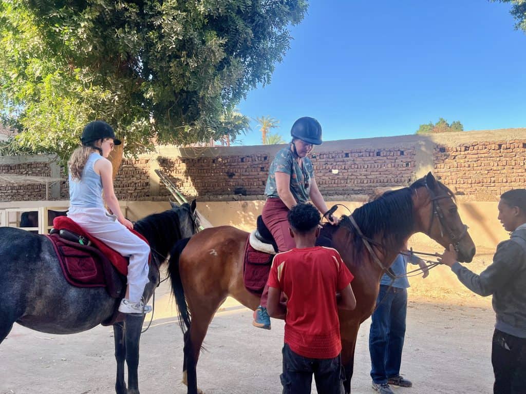 A mother and teenage daughter on horseback on a backroad while a stablehand holds the horse and another helps to adjust the stirrups.