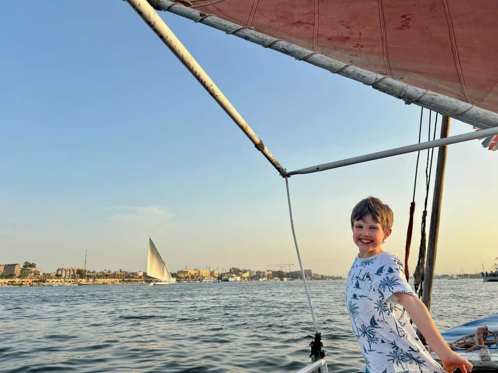 A child having a felucca ride in Luxor, Egypt. The child stands on the felucca, with the felucca sail visible above his head. In the background another felucca can be seen sailing on the river.