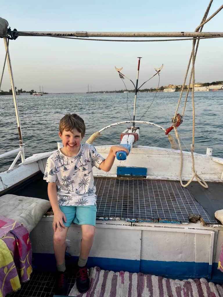 A child with his hand on the tiller of a felucca on the Nile, taking a turn at steering. In the background river cruise boats can be seen moored to the riverbank in Luxor.