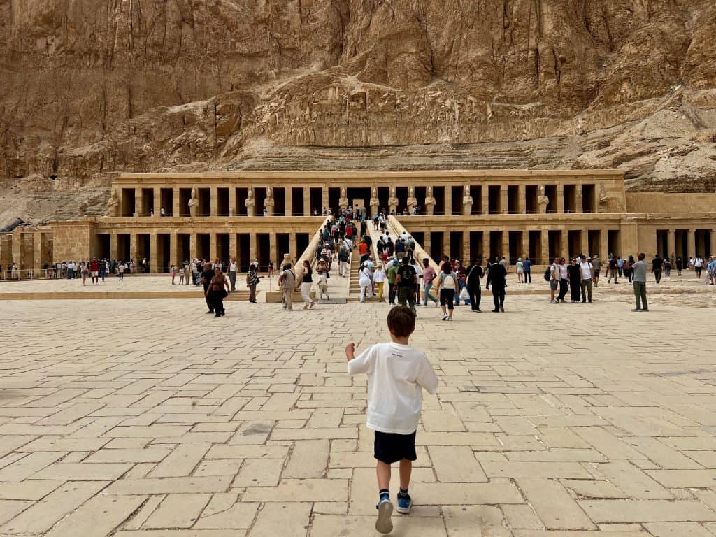 A child walking towards the temple of Hatsheput on the West Bank of Luxor, Egypt. The temple is carved into the rocks and there are lots of tourists on the steps and in front of the temple.