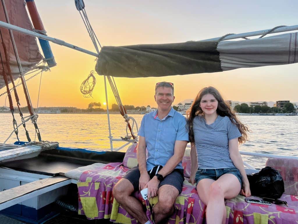 A man and his teenage daughter sit on a felucca on the Nile. Above them is the sail and in the background the sky is orange as the sun sets over the horizon.