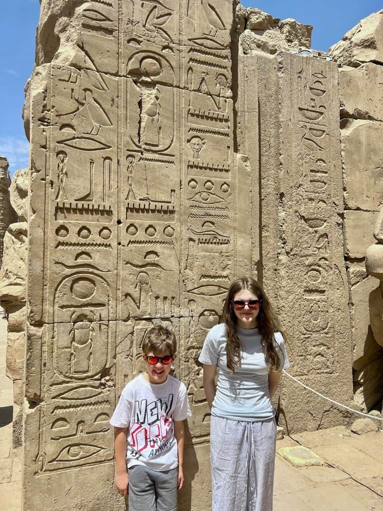 Two children stood in front of a slab of stone that is covered in Hieroglyhics, in the Karnak temple complex, Luxor, Egypt.