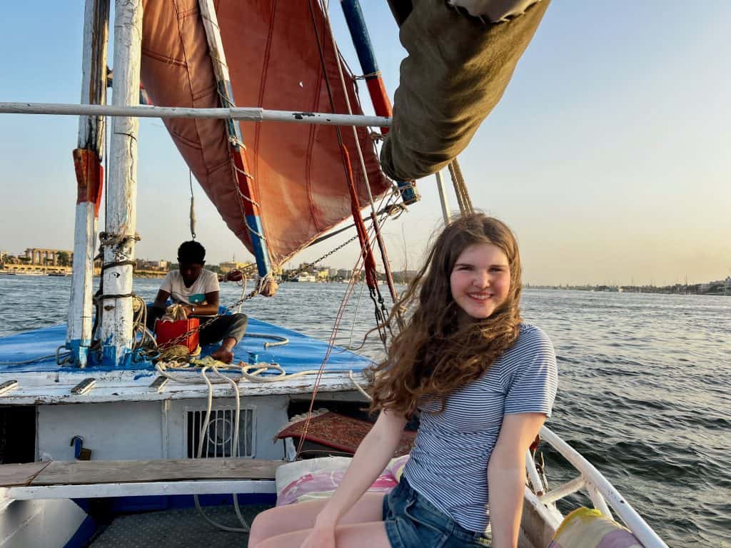 A teenager sits on a felucca on the Nile in Luxor. In the background an Egyptian boy can be seen cleaning the boat, sitting next to the mast with the sail near him.