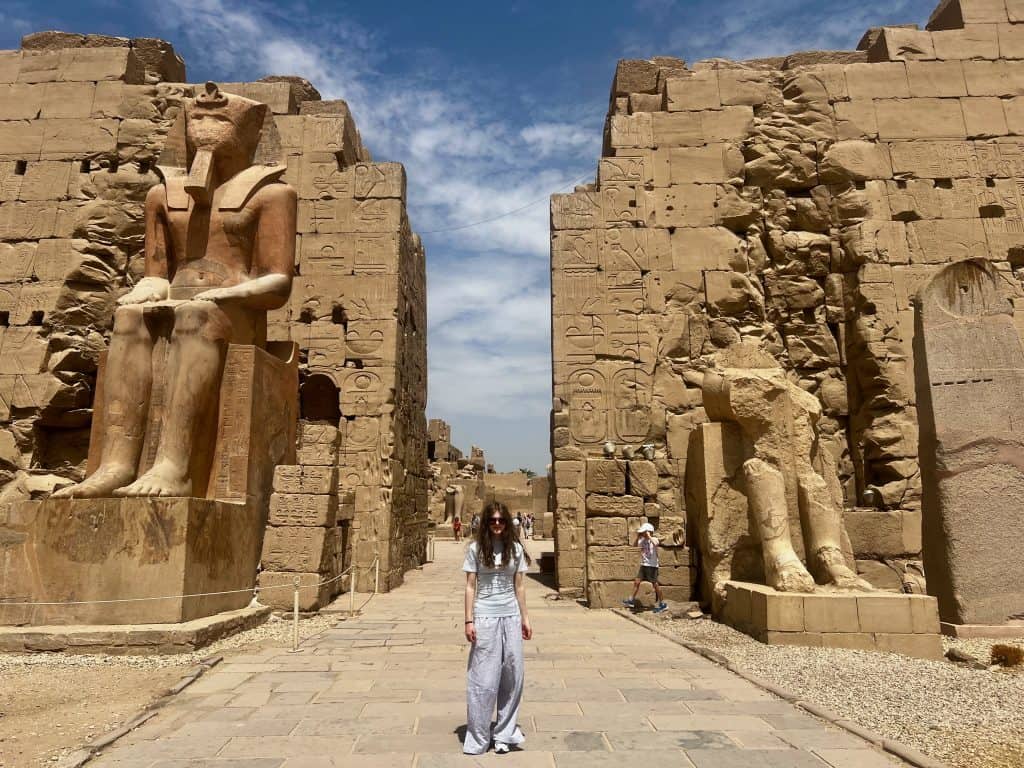 A teenager standing by the entrance to Karnak Temple in Luxor, Egypt. At either side of the large gap in the walls are statues of seated pharohoes, one of which is complete and the other has only the legs remaining.