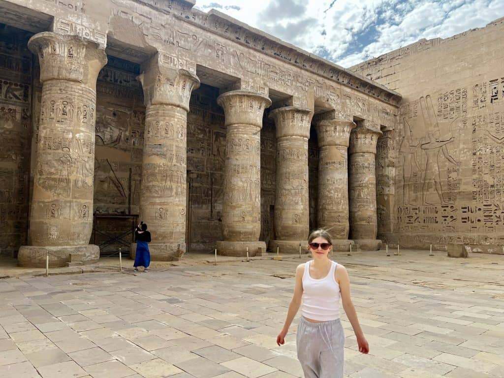 Chloe standing in the central part of Habu temple on the West Bank of Luxor, Egypt. It is open air with a line of large columns covered in hieroglyphics.