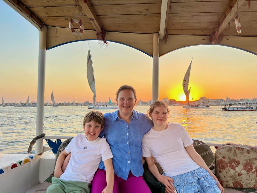 Mother and two children on a boat on the Nile in Luxor. In the background are feluccas sailing by at sunset.