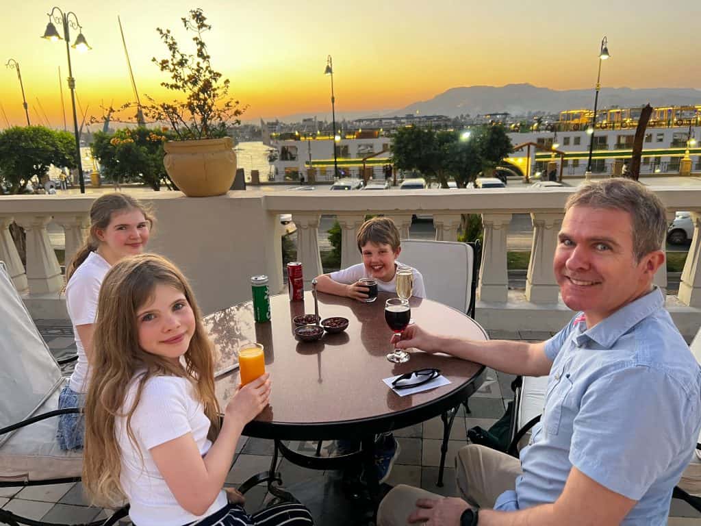 A family having drinks on the terrace of the Winter Palace hotel in Luxor. In the background there is a river cruise ship on the Nile and the sun is setting behind the mountains on the far bank of the river.