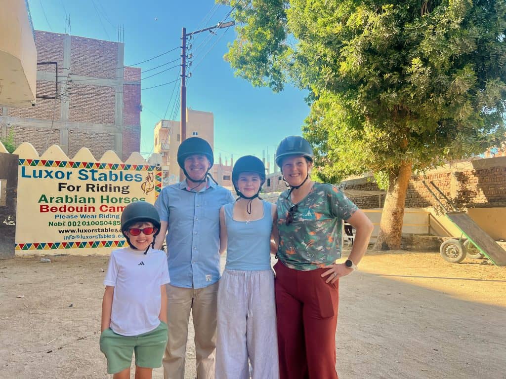 A family standing with riding helmets on in front of the sign for Luxor stables. They are standing on a dirt road with buildings and a large tree visible in the background. 