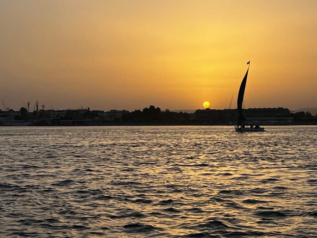 A felucca sailing on the river Nile, with a yellow sky and the sun setting beyond the horizon in the background.