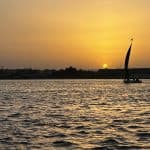 A felucca sailing on the river Nile, with a yellow sky and the sun setting beyond the horizon in the background.