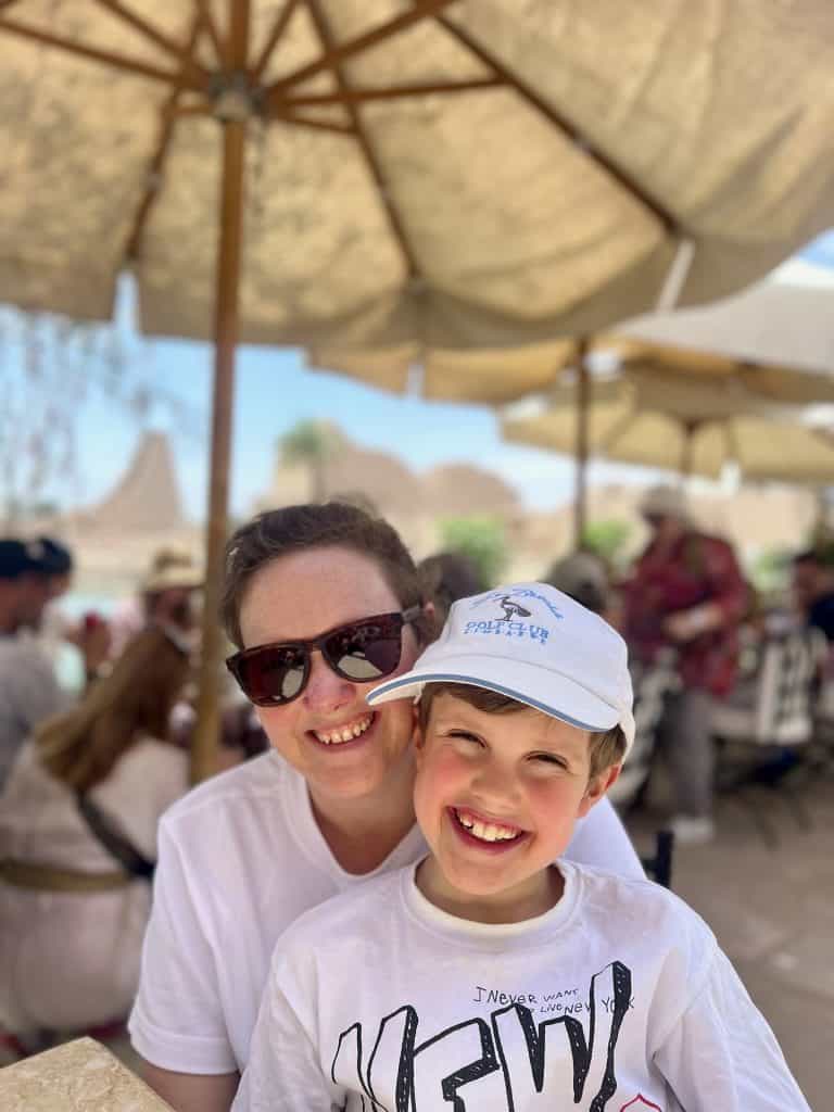Mother and son smiling while sitting in a cafe. In the blurred background can be seen people sat at other tables and beyond that the sacred lake of Karnak Temple in Luxor.