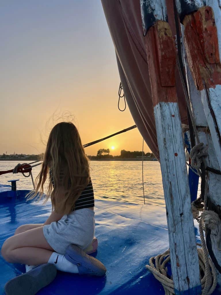 A child sitting near the prow of a felucca sailing on the Nile in Luxor. The sun can be seen setting behind the west bank in the distance.