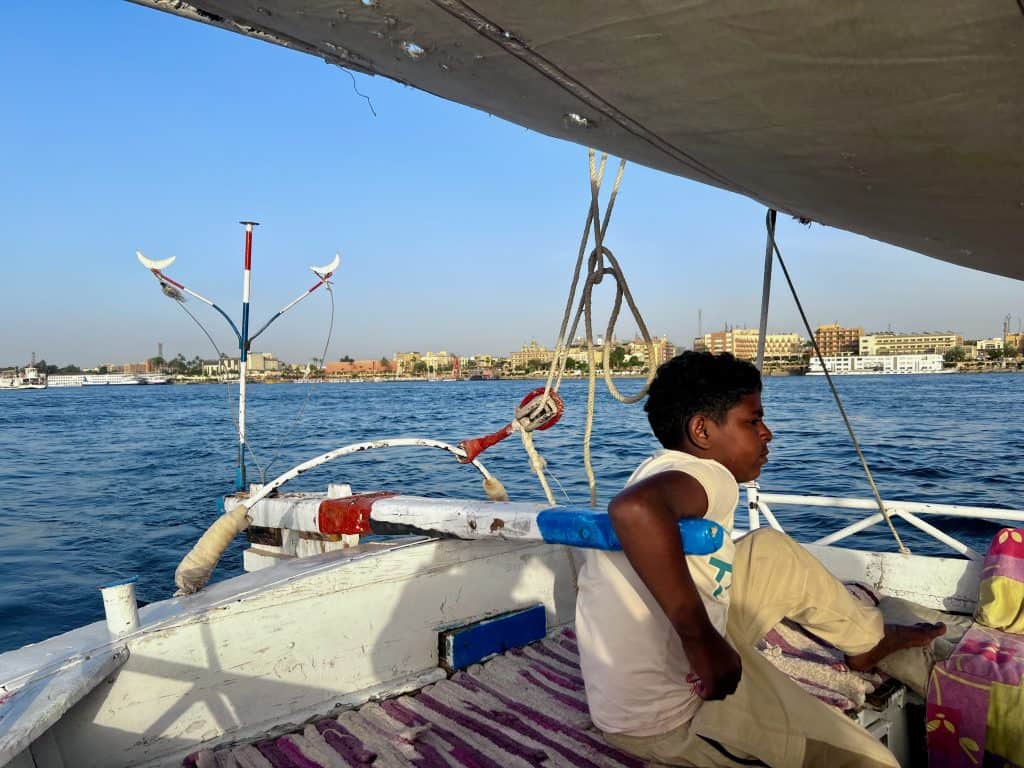 An Egyptian boy has his arm over the tiller steering a felucca on the Nile. In the background, buildings of the city of Luxor can be seen on the far river bank.