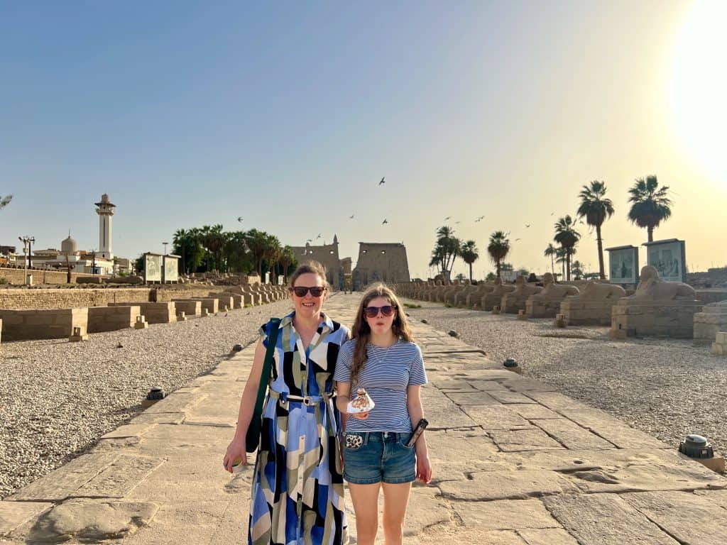 Chloe and I standing on avenue of the sphinxes in Luxor with Luxor temple visible in the background. It is a paved road flanked by sphinx statues. In the background to the left is the minaret of a mosque and on the right are palm trees.