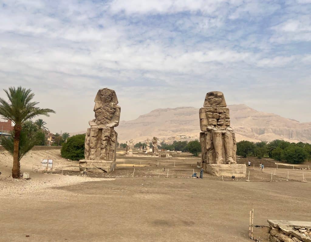 View of the Colossi of Memnon on the West Bank of Luxor, Egypt. Two large statues can be seen in the foreground with other statues in the distance and mountains visible in the background.