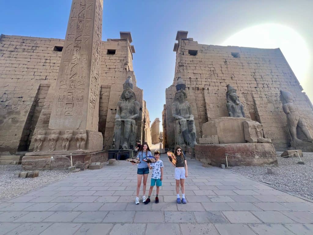 Three children showing off their newly bought fans at the entrance to Luxor temple. A large obelisk is on the right with the entrance flanked by large statues with tall walls behind them.