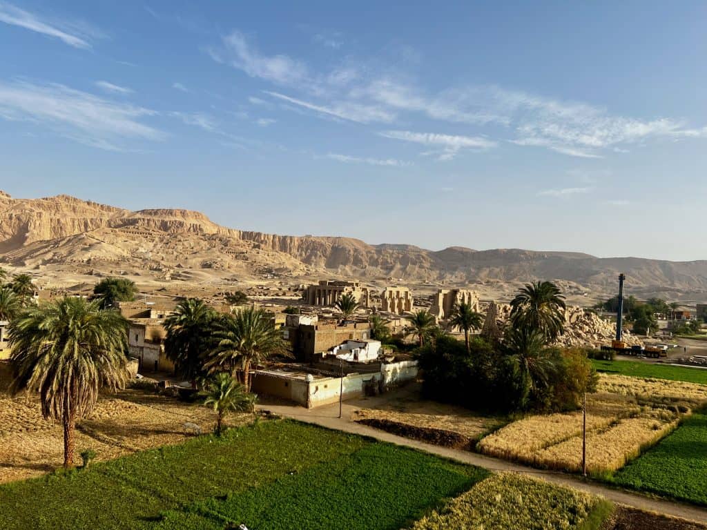 An aeriel view of the outskirts of Luxor, Egypt. In the foreground are green fields and palm trees. Behind this the ruins of an ancient temple can be seen with mountains in the background.