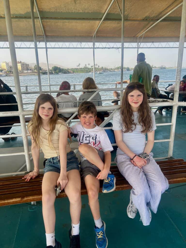 Three children sat on a wooden bench on the top deck of the Luxor River ferry crossing the Nile.