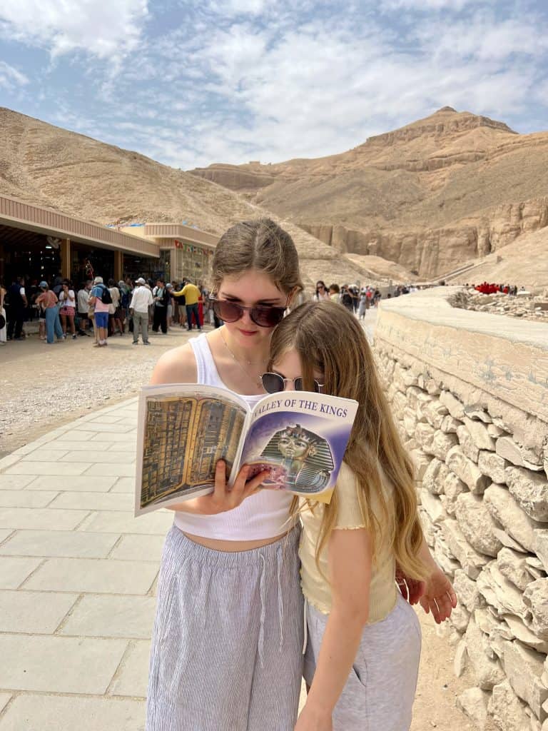Two girls reading a book about Valley of the Kings. Behind them a crowd of people are in front of the shop/ snack bar and in the distance the hills that contain the tombs of the Valley of the Kings.