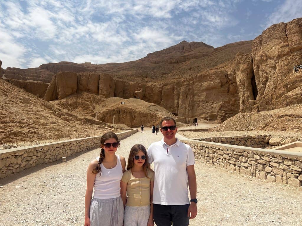 Dad and two daughters standing in valley of the kings, Luxor. Behind a dirt road can be seen reaching towards the cliffs of the mountains.