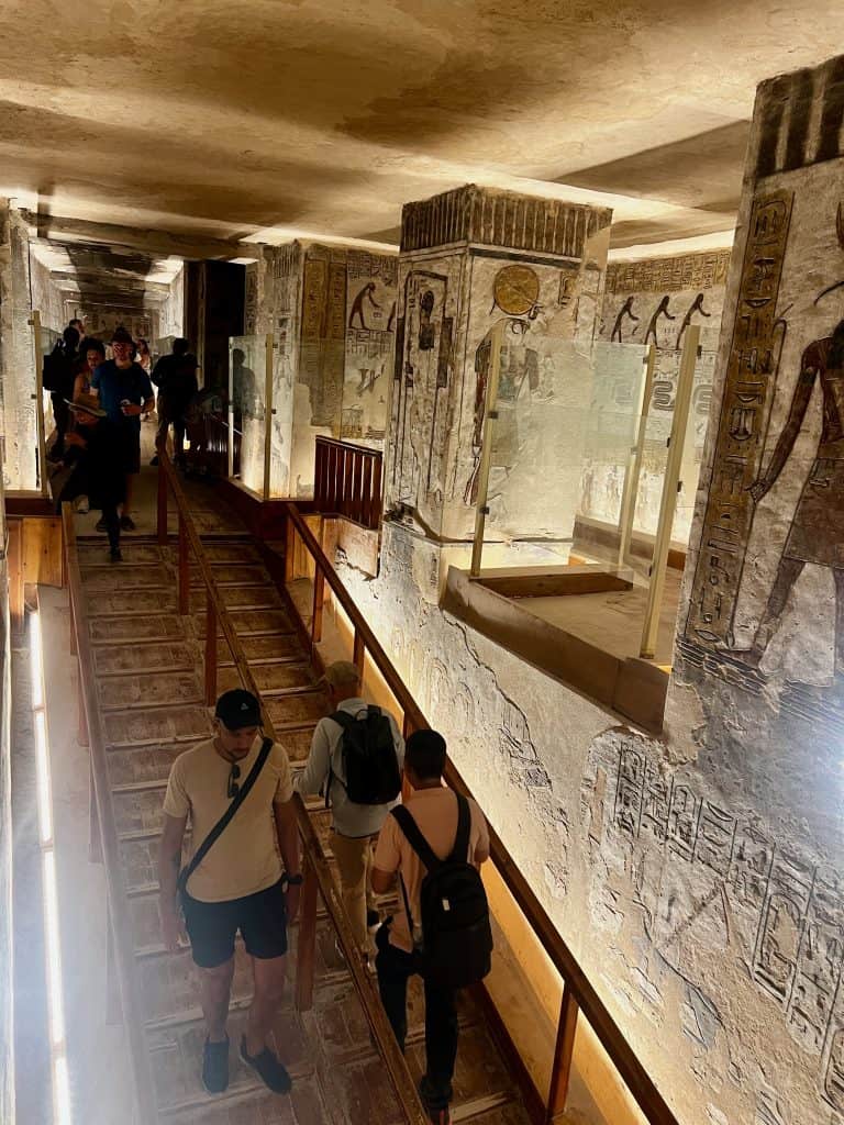 A view inside the tomb of Ramases III in valley of the kings, Luxor. People can be seen walking up and down a wooden ramp with walls containing egyptian decoration to the right, with alcoves with balconies containing glass protection.