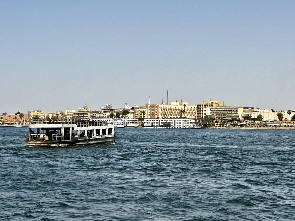 View of the river Nile in Luxor. The river ferry can be seen from behind crossing to the East Bank, with moored river cruise ships and buildings on the East bank visible in the distance.
