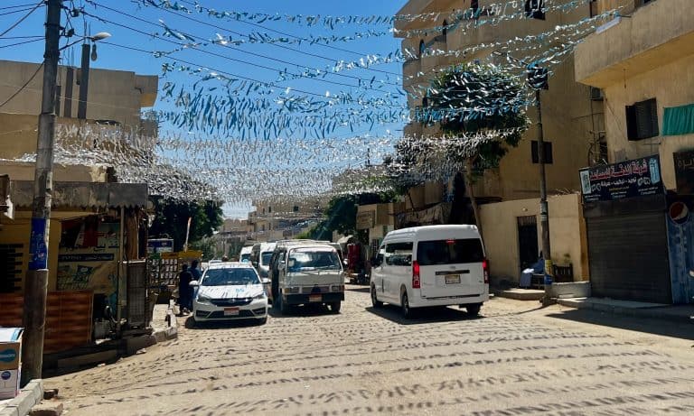 A street view in Luxor, showing cars and minivans driving down the road with buildings on either side.