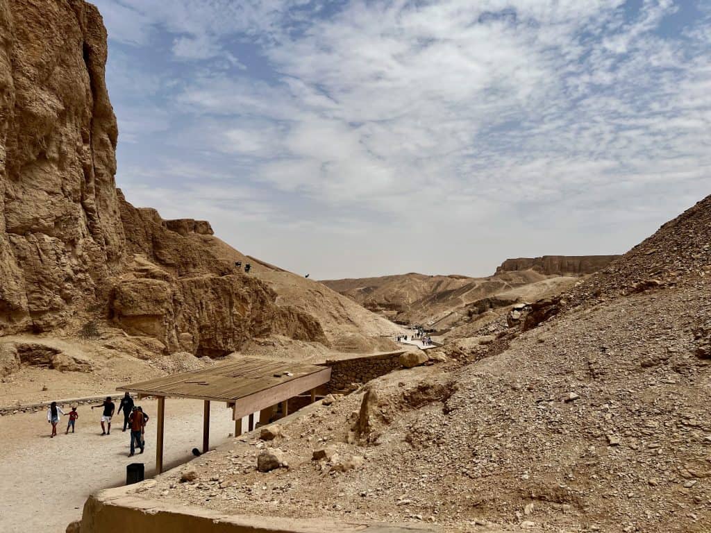 View down Valley of the Kings from near the entrance to KV14. The dirt road can be seen with brown, rocky mountains to the side. Wooden shelters are by the side of the dirt road.
