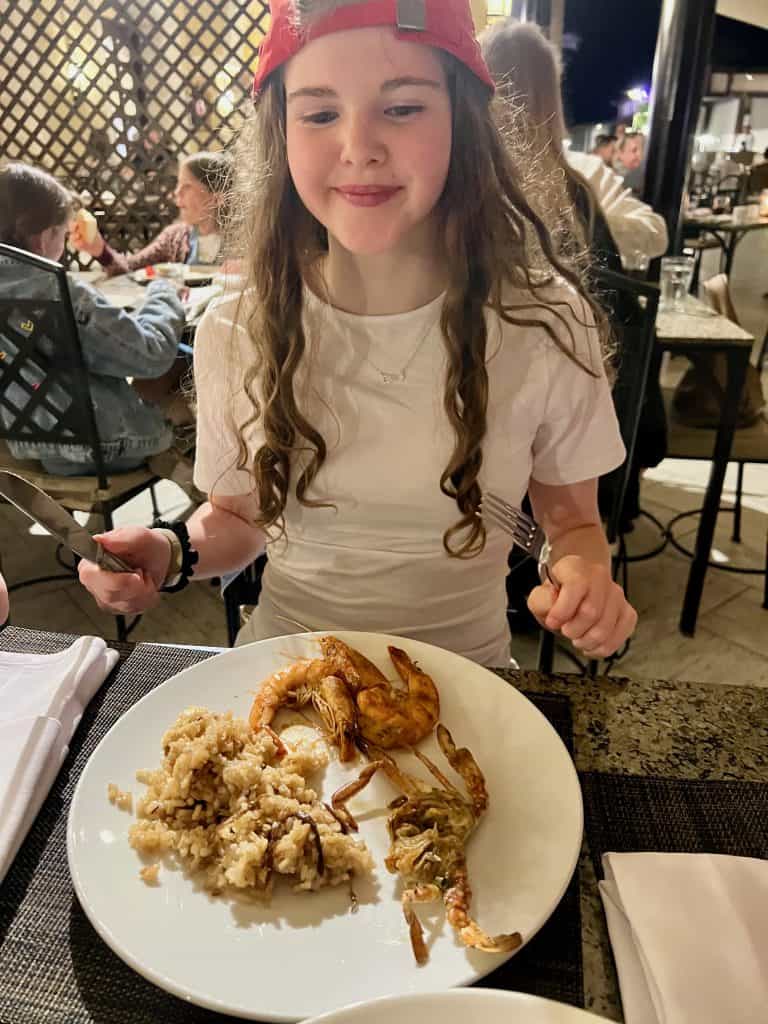 Teenager sits at a table holding a knife and fork. In front of her on her plate is a soft shell crab, some large prawns and rice.