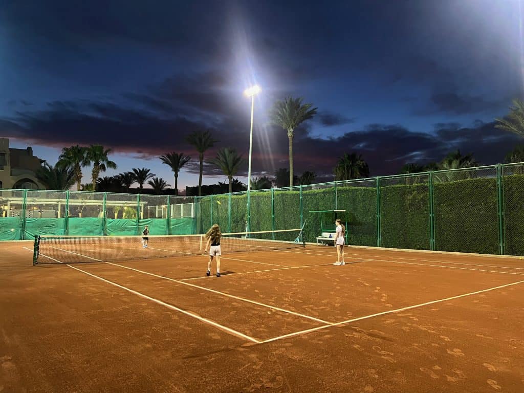 Children playing tennis on a floodlit tennis court. the sky is dark with a few clouds and palm trees are visible in the background.