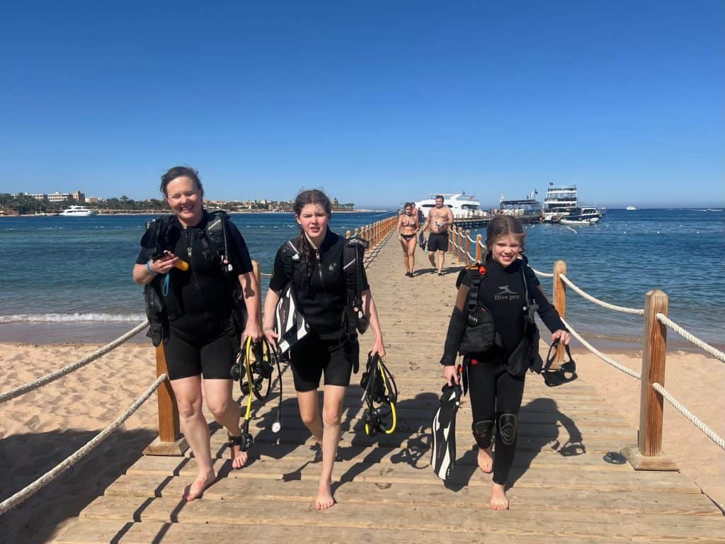 Mum and two daugthers walking down a jetty wearing wetsuits and diving BCDs, in Makadi bay near Hurghada in Egypt.