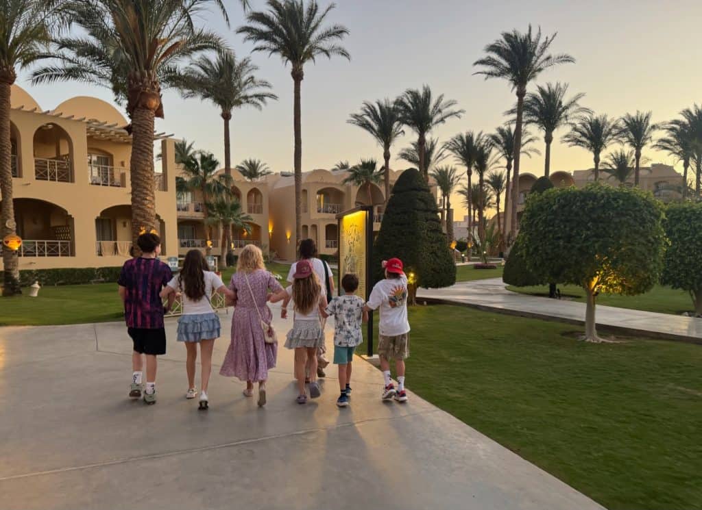 Family viewed from behin walking through the garden area of Jaz Makadina hotel in Makadin Bay. Lots of palm trees are visible and 2-storey blocks of hotel bedrooms.