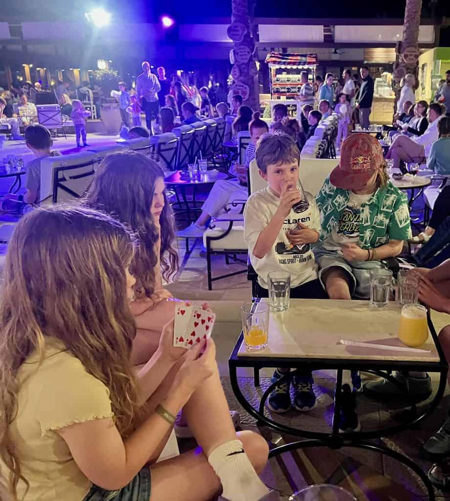 In the front of the photo is a group of children, sat around a table with drinks, playing cards. In the background are rows of other seats in the outdoor bar at Jaz Makadina