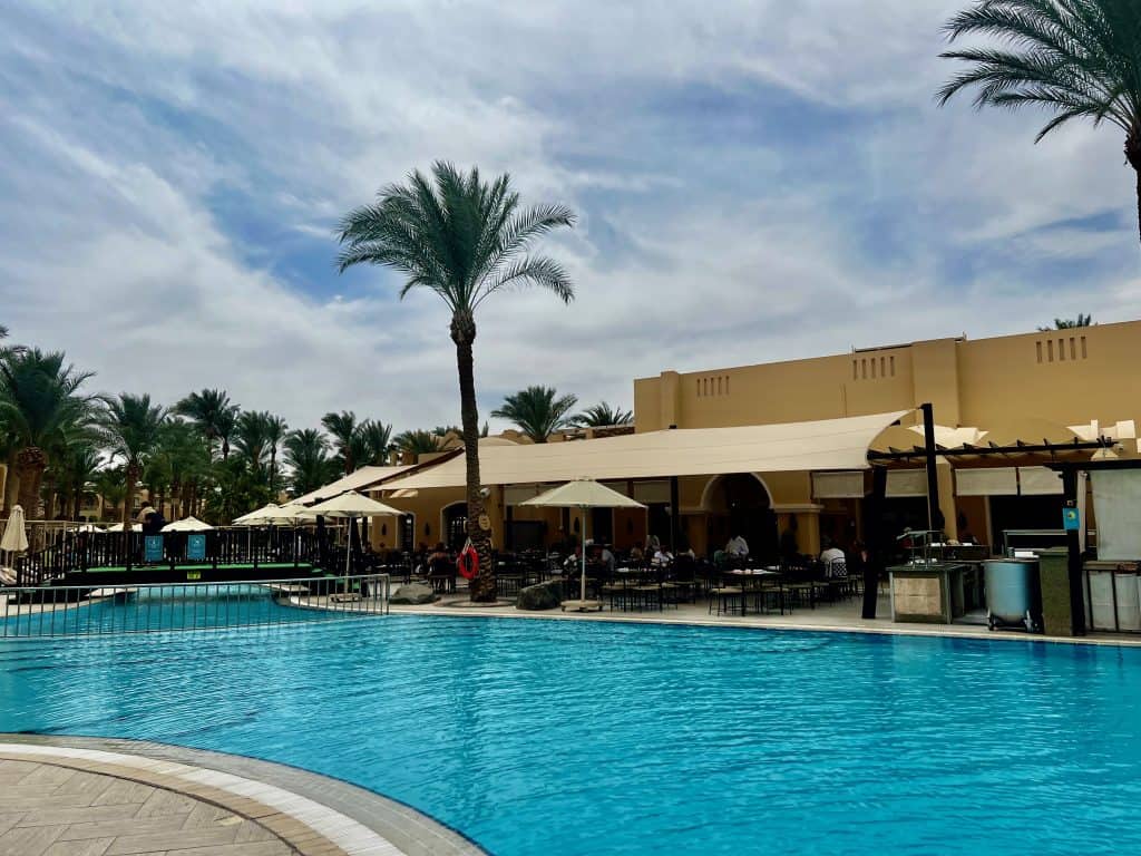 In the foreground is the hotel swimming pool and behind this tables for the restaurant at Jaz Makadina hotel are visible. To the left of the picture are palm trees.