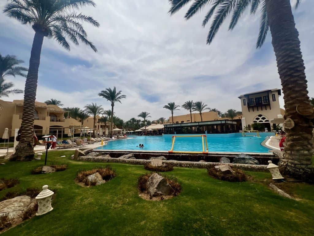 View of the swimming pool at Jaz Makadina hotel, palm trees are dotted around, in the foreground is a grassy area and the hotel restaurant and buildings are in the background.