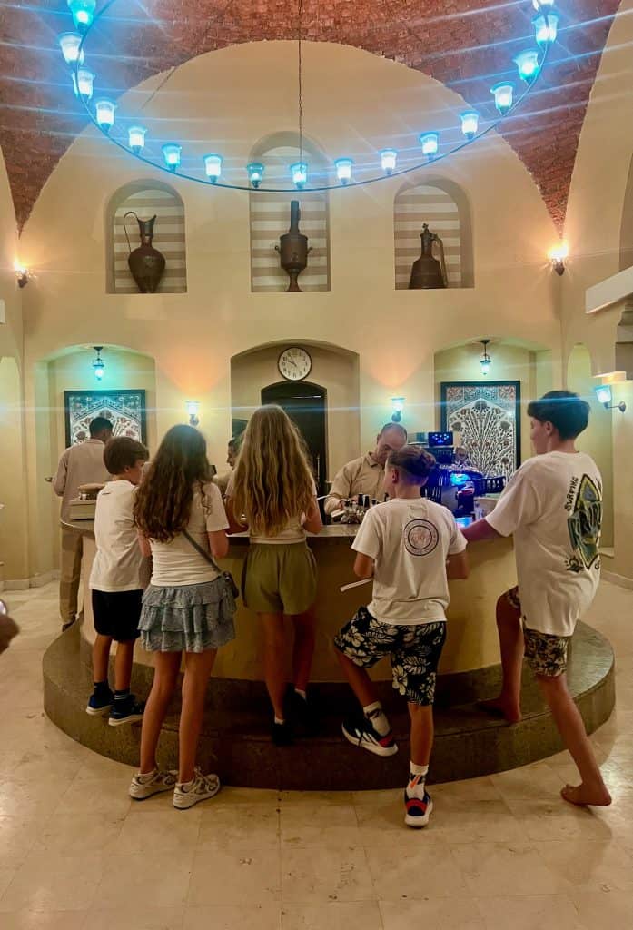 A group of tweens and teenagers stood around a curved bar while the bartender makes mocktails. Above the bar, on the far wall are recesses with large brass vases and a circle of lights hangs above shining down from the vaulted ceiling.