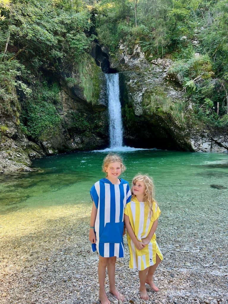 Two girls stood wearing Dock and Bay towel ponchos in front of a waterfall falling into a clear green pool of water.