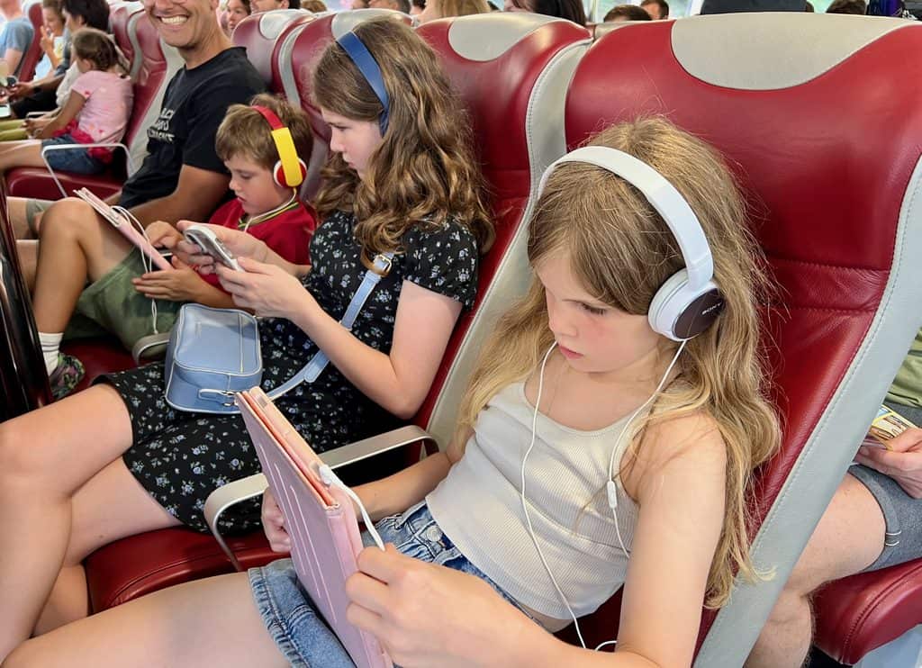 Three children sat on the Vienna- Bratislava river ferry with headphones on and looking at electronic devices.