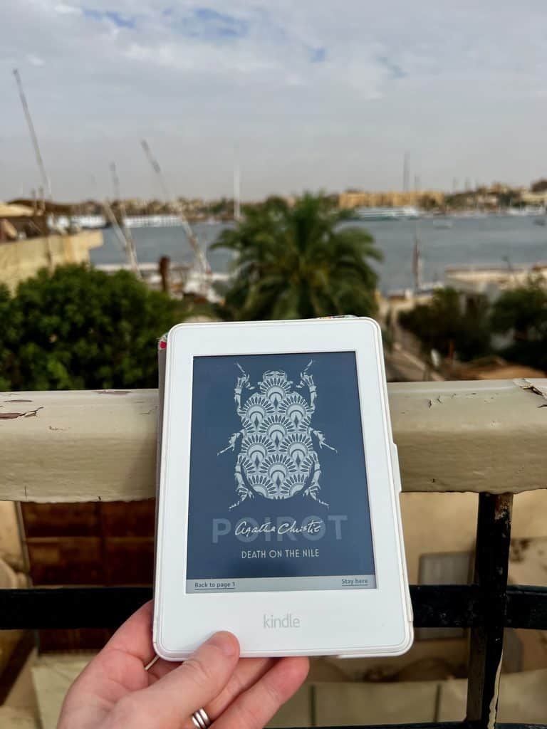 An paperwhite kindle showing the coverpage of Death on the Nile held in a hand on the edge of a balcony. In the background are palm trees, buildings and the river Nile.