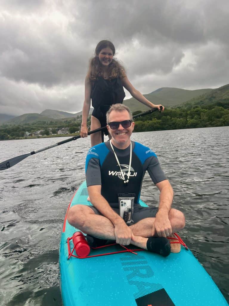 Man sat on paddleboard with teen girl stood behind him holding the paddle on Llyn Padarn in North Wales.