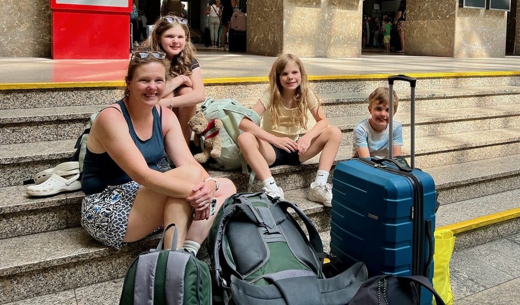 A family sit on the steps at Bratislava train station with a suitcase and several backpacks around them on the floor.